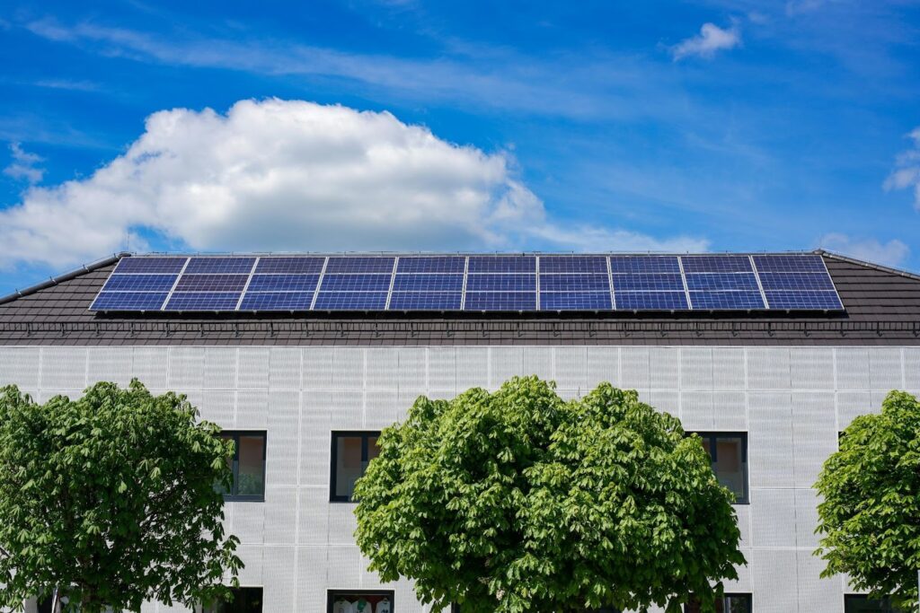 大片太陽能電池板陣列安裝在戶外的草地或綠色空間上，背景是工廠或倉庫等工業建築，藍天白雲襯托著，象徵著綠色能源、太陽能發電、環保科技或企業對永續發展的承諾。